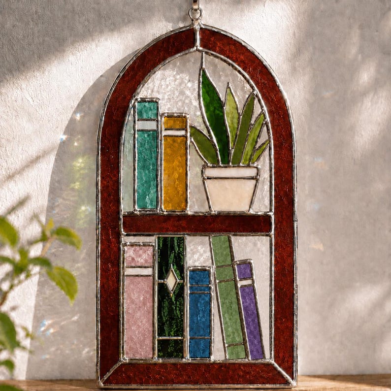 Puede incluir: Estanter&iacute;a de vidrieras con una planta en maceta y libros coloridos. El marco es de color marr&oacute;n rojizo oscuro, y los libros son de varios colores, incluyendo verde, amarillo y azul. La planta tiene hojas verdes y est&aacute; en una maceta blanca.