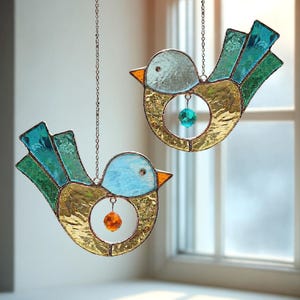 Könnte beinhalten: Zwei Glasmalerei-Vogelornamente, die an Ketten hängen. Die Vögel haben blaue Köpfe, goldene Körper und türkisfarbene Flügel. Jeder Vogel hat eine kleine, runde, juwelenfarbene Perle, die in der Mitte seines Körpers hängt. Die Vögel hängen vor einem Fenster.