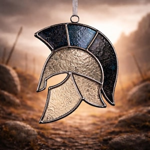 May include: Stained glass Spartan helmet ornament with a silver-colored metal frame. The helmet features sections of blue, black, and gold-colored glass. A white ribbon is attached for hanging. The background is a blurred image of a battlefield.