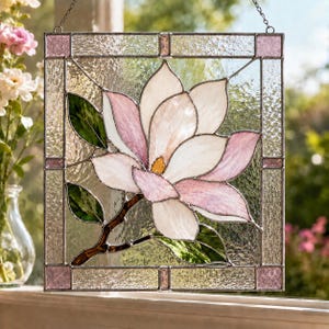 Op de afbeelding: Een vierkante glas-in-lood raamhanger met een centrale magnolia bloem met witte en zachtroze bloemblaadjes, een geel hart en groene bladeren. Het paneel is omlijst door een metalen rand met kleine roze glazen vierkantjes op de hoeken.
