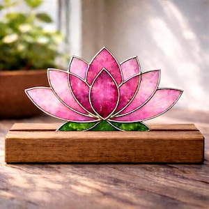 May include: A pink stained glass lotus flower with silver-colored metal outlines, set on a wooden base. The lotus has a green base and is displayed in front of a blurred background.