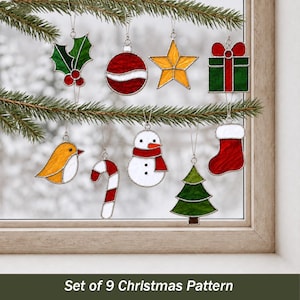 May include: A set of nine stained glass Christmas ornaments featuring a holly leaf, red bauble, yellow star, green gift, yellow bird, candy cane, snowman, green tree, and red stocking, all hanging from pine branches with silver metal borders.