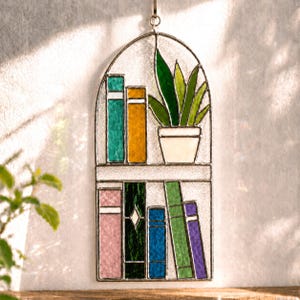 Puede incluir: Estanter&iacute;a de vidrieras con libros coloridos y una planta en maceta. El dise&ntilde;o presenta un marco de metal y una variedad de colores, incluyendo verde, azul, naranja y morado. La planta est&aacute; en una maceta blanca.