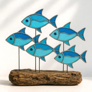 Puede incluir: Una escultura decorativa con cinco peces de vidrieras azules montados sobre varillas met&aacute;licas individuales, colocados en un trozo de madera flotante. Los peces tienen diferentes tonos de azul y est&aacute;n acentuados con ojos azul oscuro y contornos plateados.