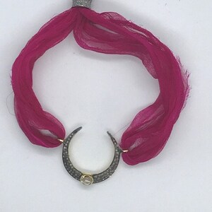May include: A magenta silk bracelet with a crescent moon pendant. The pendant is encrusted with small, dark stones and a central gold-colored gem. The bracelet is tied with a silver-colored band.