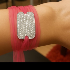 May include: A silver bracelet featuring a rectangular, gem-covered centrepiece. The bracelet is wrapped in a sheer, bright pink fabric. The jewellery is a fashion accessory.