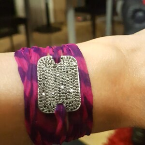 May include: A striking bracelet with a rectangular silver-toned clasp covered in small, dark stones. The band is made of flowing fabric in pink and purple, creating a bold contrast. The bracelet is designed to be worn around the wrist.