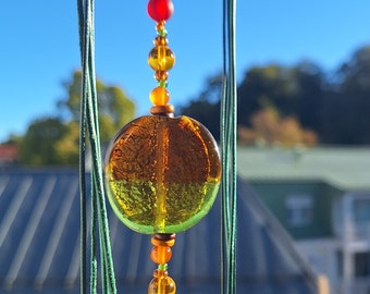 May include: A glass bead with a green and amber color gradient hangs from a string of colorful beads. The beads are red, orange, yellow, and green. The background is out of focus and shows a blue sky and green trees.