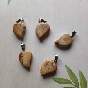 May include: Five heart-shaped pendants made of tan and brown stone with silver-colored metal bails. The pendants have unique patterns and textures, with variations in color and veining. The pendants are arranged on a light-colored surface.