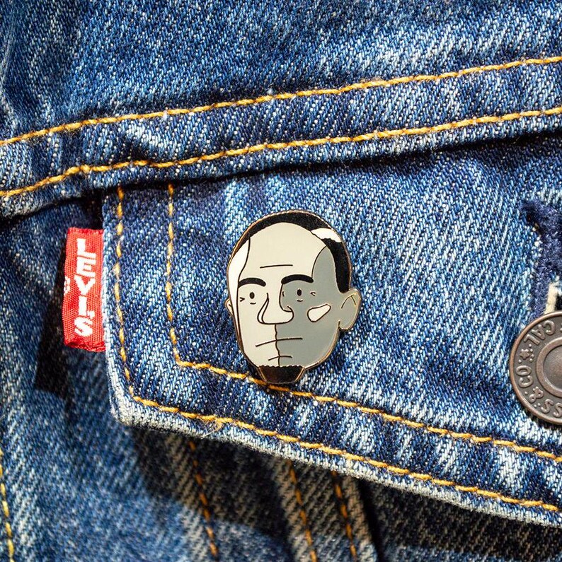 May include: A gold-colored enamel pin featuring a black and white illustration of a person's face. The pin is attached to a denim jacket.