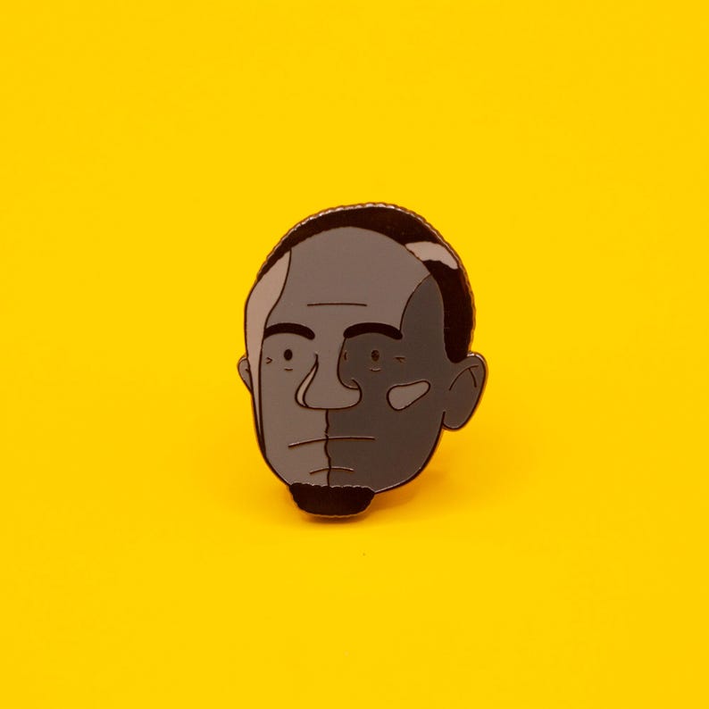 May include: A black and white enamel pin of a person's face. The pin is a cartoon style illustration of a person with a black goatee and a black hairline.