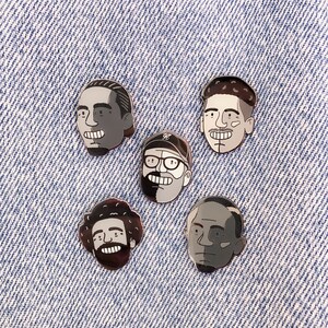 May include: Five black and white enamel pins featuring different cartoon faces. The pins are on a blue denim background.