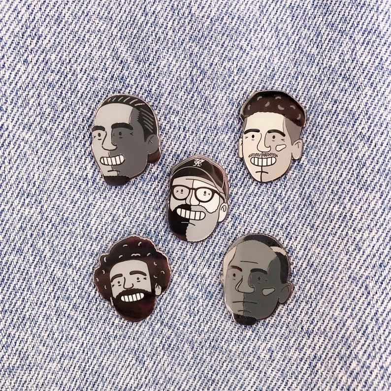 May include: Five black and white enamel pins featuring different cartoon faces. The pins are on a blue denim background.