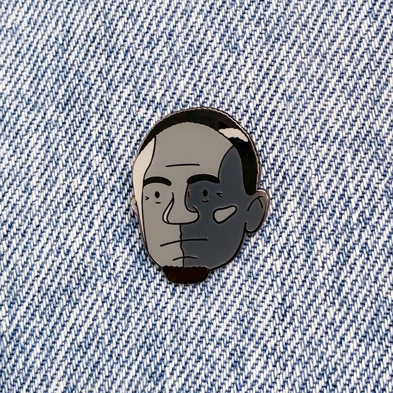 May include: Black and white enamel pin of a person's face. The face is split down the middle, with one side black and the other white.