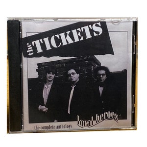 May include: A CD case with a black and white photo of three men on the front. The band's name, "The Tickets", is written in large, bold letters on a torn piece of paper. The text "the complete anthology" and "local heroes" are also visible on the front of the case.