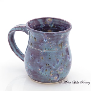 May include: A handmade ceramic mug with a blue and purple glaze. The mug has a rounded body, a sturdy handle, and a slightly flared rim. The glaze has a speckled appearance with hints of gold. The mug is suitable for hot or cold beverages.