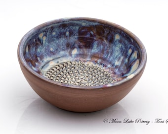 Pode incluir: Uma tigela de cerâmica artesanal com interior texturizado e borda esmaltada azul e roxa iridescente. O exterior é de cor castanha quente. O interior da tigela apresenta uma superfície texturizada e padronizada. Perfeita para servir ou expor.