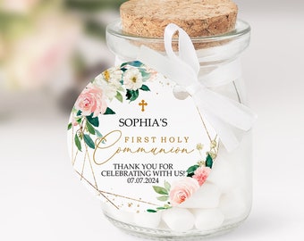May include: A small glass jar with a cork stopper, filled with white candies. A white ribbon is tied around the neck of the jar, holding a round tag with floral and gold geometric designs. The tag reads "Sophia's First Holy Communion".