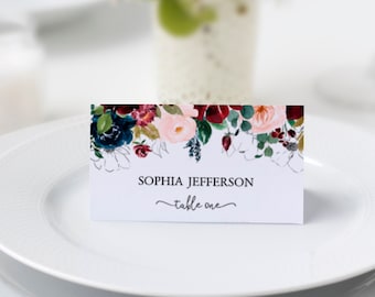 May include: A white place card with a floral design in shades of red, pink, blue, and green. The text on the card reads "Sophia Jefferson - table one".