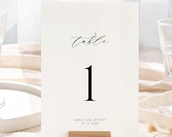 May include: White table number card with black calligraphy that reads "Table 1" and the text "Amely and Jeffery Oct. 11, 2024". The card is on a wooden stand.
