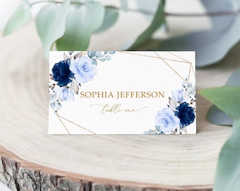 May include: A white place card with a gold geometric frame and blue watercolor flowers. The text on the card reads "Sophia Jefferson, table one."