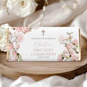 Puede incluir: Envoltorio rectangular de barra de chocolate con rosas rosas y una cruz. El texto dice: "Primera Comunión de Christina, 23 de junio de 2027". Sobre una bandeja de madera con flores blancas.