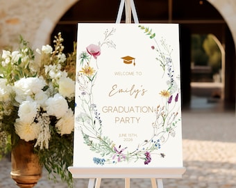 May include: A white welcome sign with a floral wreath border and gold text. The sign reads "Welcome to Emily's Graduation Party June 15th, 2026".