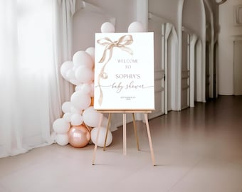 May include: A welcome sign for a baby shower, displayed on a wooden easel. The sign features a gold ribbon design and the words "Welcome to Sophia's baby shower." White and rose gold balloons decorate the background.