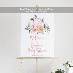 May include: A white welcome sign with pink floral accents and two pink pumpkins. The sign reads "Welcome to Sophia's Baby Shower October 15th, 2019".