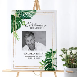 May include: A white memorial sign with a gold frame and green tropical leaves. The sign says "Celebrating the Life of" and includes a black and white photo of a man. The text below the photo reads "Andrew Smith - September 22, 1960 - July 15, 2030".