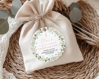 Könnte beinhalten: Ein runder, weißer Geschenkanhänger mit einem floralen Kranzdesign. Der Anhänger trägt die Aufschrift "Olivia's First Holy Communion" in rosa Schrift mit einem Kreuz und dem Datum "07.07.2026". Der Anhänger ist an einem kleinen, weißen Stoffbeutel befestigt.