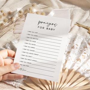 May include: A white card with black text that reads "prayers for baby" and includes a list of prayers for a baby. The card is held in a hand.