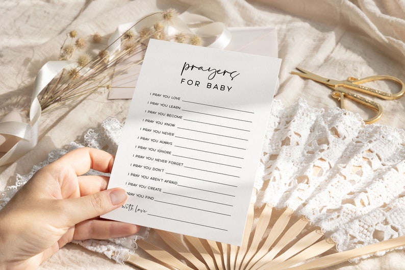 May include: A white card with black text that reads "prayers for baby" and includes a list of prayers for a baby. The card is held in a hand.