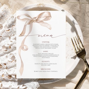 May include: A wedding menu card featuring a watercolour bow in beige and brown tones. The menu lists starters, mains, desserts, and a mimosa bar. The card is on a white plate with gold cutlery.
