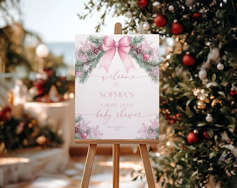 Pode incluir: Uma placa de boas-vindas para chá de bebê em um cavalete de madeira. A placa apresenta um laço rosa, flocos de neve e o texto "welcome to Sophia's, A Merry Little Baby Shower, December 16, 2026". A placa é decorada com vegetação e ornamentos festivos.