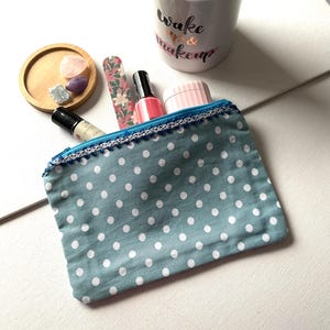 May include: A teal polka dot makeup bag with a blue zip and white trim, containing nail polish, a nail file, and other beauty products. A ceramic cup with the words "wake up & makeup" is in the background.