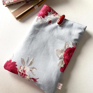 May include: A pale blue fabric book cover featuring a red and white floral design, held closed by a red clip. A stack of notebooks with a matching floral pattern is in the background.