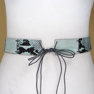 Op de afbeelding: Een lichtblauwe en zwarte slangenleren riem met een strik sluiting. De riem is gemaakt van leer en heeft een uniek ontwerp.