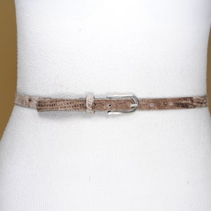 Può includere: Una cintura sottile, marrone chiaro, con una trama simile a quella di un rettile è allacciata attorno a un manichino bianco. La cintura ha una fibbia color argento. È un accessorio di moda.