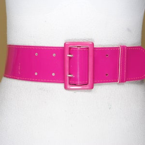 Pode incluir: Um cinto largo rosa brilhante com um acabamento brilhante. O cinto apresenta uma fivela retangular e uma presilha a condizer. O cinto é mostrado em um manequim branco, destacando sua cor e design arrojados. O cinto tem cerca de 7,6 cm de largura.