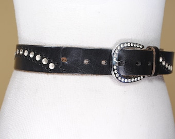 May include: A black leather belt with a silver buckle and silver studs. The buckle is a horseshoe shape with rhinestones.