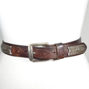 May include: A brown leather belt with silver-toned metal accents. The belt features a silver buckle and decorative rectangular plates. The belt is worn around a white mannequin form.