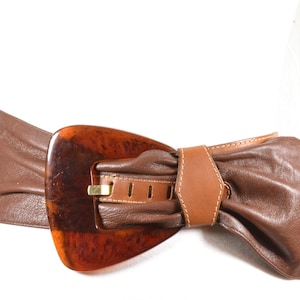 Peut inclure: Une ceinture en cuir marron avec une grande boucle en écaille de tortue. La ceinture est nouée en un nœud à l'avant.
