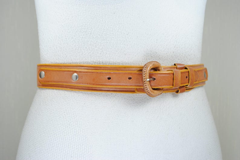 A tan leather belt with a buckle, displayed on a mannequin torso against a white background.