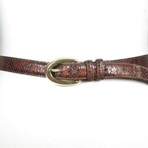 Peut inclure: Une ceinture en cuir marron avec une boucle ovale dorée. La ceinture a un motif de peau de serpent.