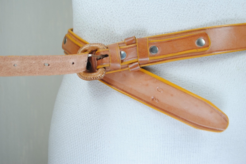 A close-up image of a tan leather belt with a metal buckle, displayed on a mannequin or mannequin head.