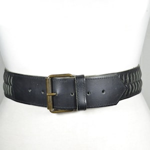 May include: Black leather belt with a brass buckle. The belt features a woven design on the top and bottom edges. The belt is displayed on a white mannequin.