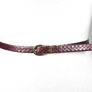 Può includere: Una cintura bordeaux con un motivo a pelle di serpente strutturato e una fibbia color oro. La cintura è mostrata su un manichino bianco, evidenziando il suo design e il colore. Un accessorio elegante.