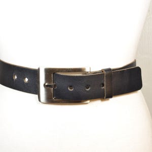 May include: A black leather belt featuring a rectangular silver-toned metal buckle. The belt has a smooth finish with visible stitching along the edges and a series of punched holes for adjustment, secured by a matching black leather keeper loop.
