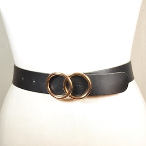 Cinturón de cuero negro con hebilla dorada de doble anilla, estilo minimalista moderno.